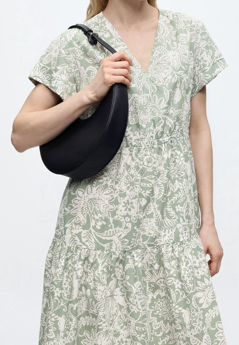 Femme portant une robe fleurie verte et blanche, tenant un sac à main en cuir noir en forme de croissant sur son épaule.
