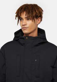 Mustapaitainen huppari, jossa on vetoketjukiinnitys, kahdella rintataskulla ja pienellä logotulostuksella. Kangas vaikuttaa sileältä ja kestävältä.