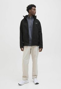 Sort softshell jakke med høj krave, grå fleece med lynlås nedenunder, cremefarvede bukser og hvide sneakers. Enkel, funktionel design.