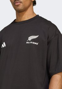 Černé bavlněné tričko s bílým logem "All Blacks" a designem kapradiny na levé hrudi, s třemi bílými pruhy na levém rukávu.