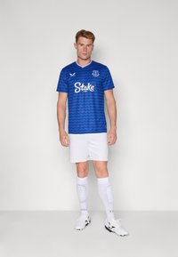 Plava nogometna majica Everton FC s uzorkom valova, bijelim akcentima i logom. Bijele kratke hlače i bijele čarape do koljena s podstavom. Crna obuća.