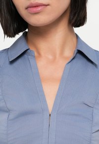 Ανοιχτό μπλε πουκάμισο με γιακά και φερμουάρ μπροστά, με λεία υφή και διακριτικές ραφές για εφαρμοστό αποτέλεσμα.