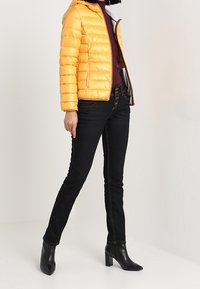 Personne portant une veste matelassée jaune, un col roulé bordeaux, un jean noir moulant et des bottines à talons noires se tenant sur un sol blanc.