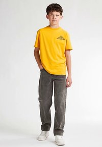 Gelbes Baumwoll-T-Shirt mit schwarzem Text auf der Brust, kombiniert mit dunkelgrauen Jeans und weißen Sneakers. Das Model steht mit den Händen in den Taschen.