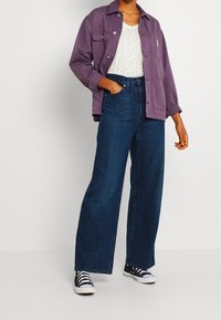 Chaqueta corta morada sobre una blusa blanca con puntos dorados, combinada con jeans anchos de tiro alto en azul y zapatillas negras.