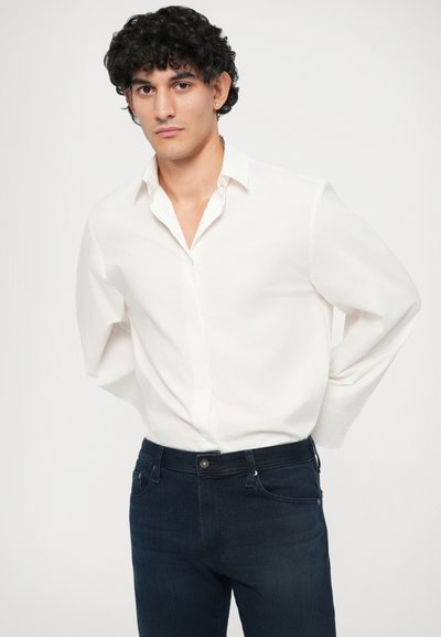 Λευκό πουκάμισο με μακριά μανίκια και γιακά σε αιχμή, συνδυασμένο με σκούρα μπλε τζιν παντελόνι. Απαλή υφή, εφαρμοστή σχεδίαση, ελάχιστες λεπτομέρειες.
