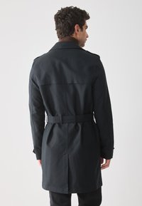 Abrigo negro tipo trench con cintura ceñida y cinturón, mangas largas y una abertura en la espalda, con un cuello marrón. Textura suave, diseño clásico.