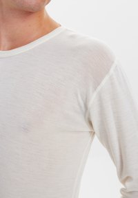 Helles cremefarbenes Langarmshirt mit subtiler Textur, Rundhalsausschnitt und kontrastierenden Nähten entlang der Kanten. Der Stoff wirkt weich und leicht.