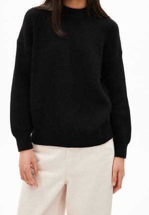 Persoon die een zwarte, gebreide trui met lange mouwen en een witte broek draagt, staand tegen een effen witte achtergrond.