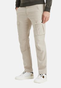 Pantalones cargo beige hechos de tela texturizada, con múltiples bolsillos y detalles de costura visibles, combinados con zapatillas blancas.