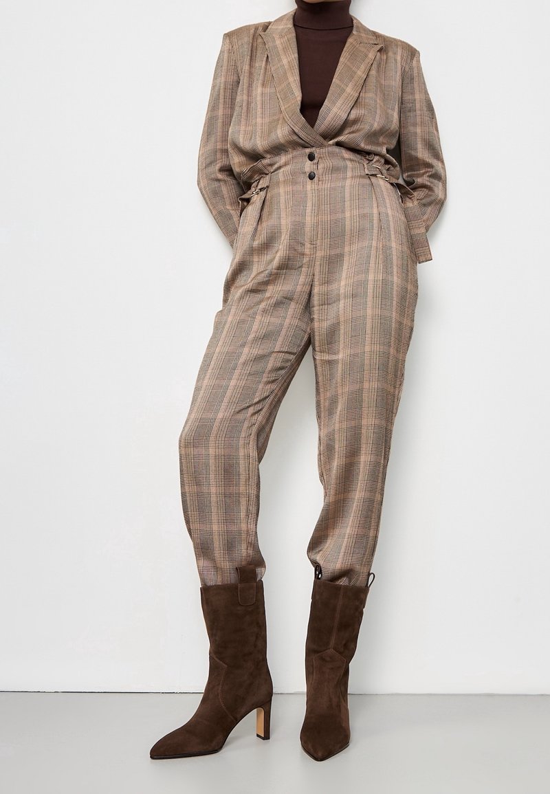 Hnědý kostkovaný oblek s dvouřadým sakem a kalhotami s vysokým pasem, zdobený dvěma knoflíky a volným střihem. Kombinováno s hnědými semišovými kotníkovými botami.