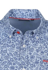 Lichtblauwe blouse met een bloemenpatroon in marineblauw. Heeft een button-down kraag, witte knopen en een rode accent nabij de kraag.