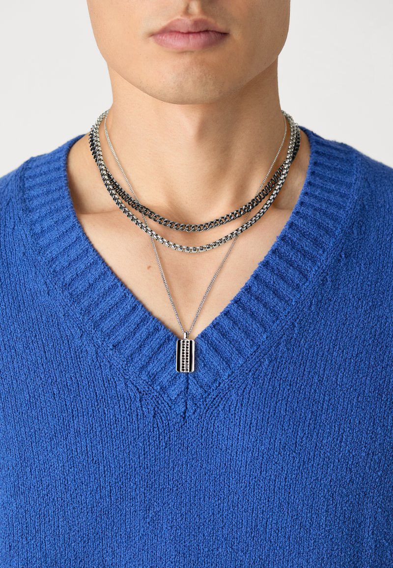 Blauwe gebreide trui met een V-hals ontwerp. Drie gelaagde metalen kettingen, waaronder één met een rechthoekige hanger, zichtbaar bij de halslijn.