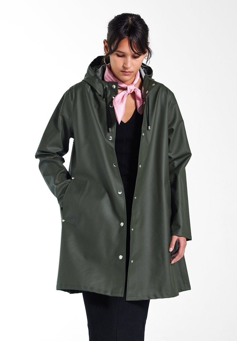 Manteau imperméable vert avec une capuche, fermetures à pression sur le devant et poches latérales. Texture lisse, coupe légèrement ample, et écharpe rose assortie.