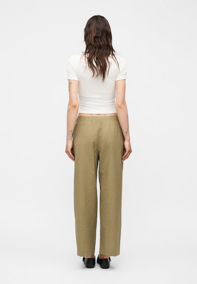 Γυναίκα με μακριά σγουρά μαλλιά, που φοράει στενό λευκό κοντομάνικο τοπ, φαρδιά ελιά πράσινα παντελόνια και μαύρα παπούτσια, στέκεται με την πλάτη προς τα εμπρός.