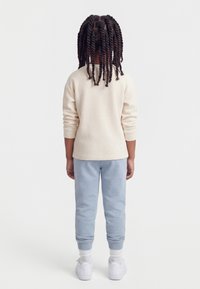 Παιδί με στριφτά κοτσίδια στέκεται γυρισμένο προς τα πίσω, φοράει κρεμ μακρυμάνικο πουκάμισο, ανοιχτό μπλε παντελόνι, λευκές κάλτσες και λευκά αθλητικά παπούτσια.