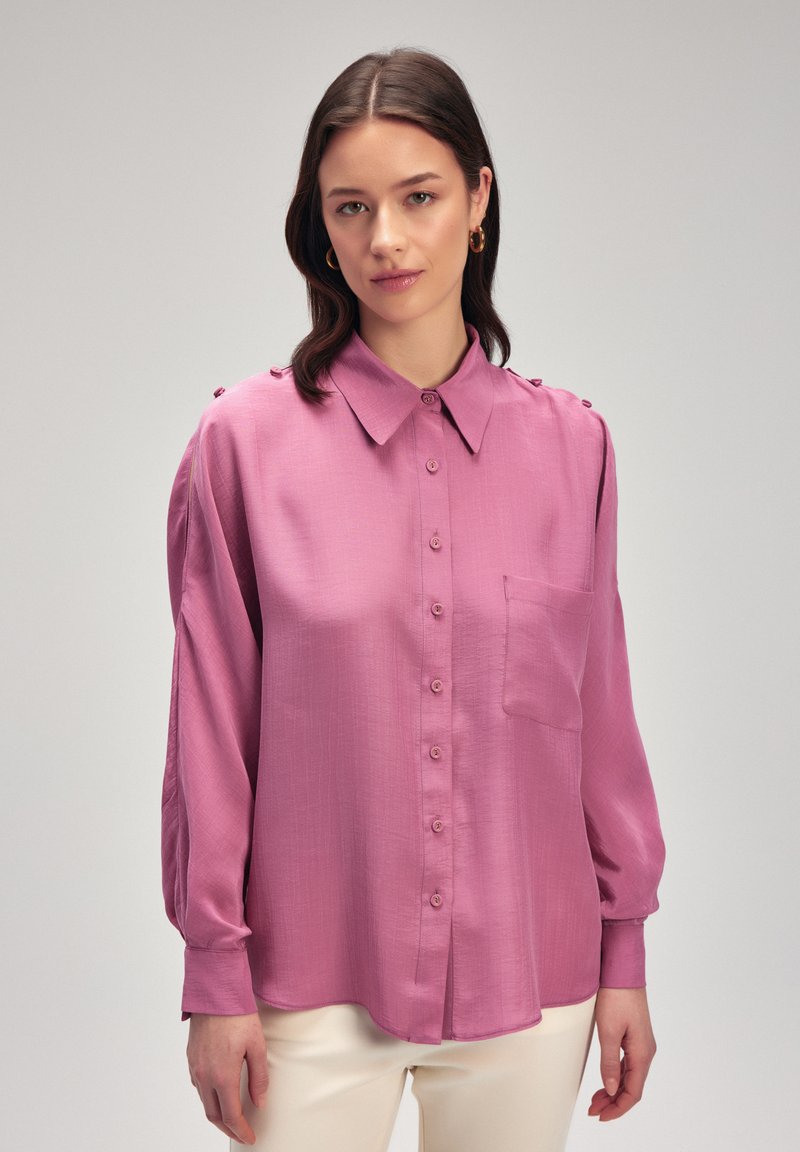Blusa color rosa con abbottonatura frontale, colletto, tasca sul petto singola e un leggero lucido; presenta maniche lunghe con dettagli a bottoni.