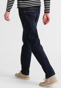 Jeans azul oscuro con un corte recto, que presentan detalles de costura sutil y un diseño clásico de 5 bolsillos, combinados con zapatos color beige claro.