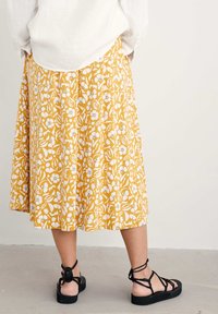 Gul kjol med vitt blommönster, plisserad design och medellång längd, ihop med svarta sandaler med remmar.