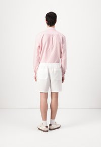 Camicia a maniche lunghe a righe rosa chiaro, pantaloni corti bianchi tessuti, scarpe bianche con accenti marroni, vista da dietro.
