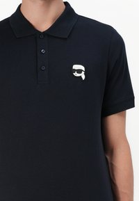 Mornarska polo majica iz bombaža, z gumbnim zapiranjem s tremi gumbi in značilnim belim ter črnim logotipom na levem prsnem delu.