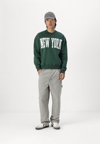Green sweatshirt with "NEW YORK" in white letters, gray pants, and white sneakers with black accents. Gray cap completes the outfit.