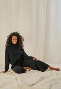 Schwarzer gerippter Pullover mit hohem Kragen, kombiniert mit lockeren schwarzen Hosen. Modell sitzt vor einem hellbeigen Stoffhintergrund.