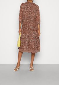 Braun geblümtes Kleid mit hohem Ausschnitt, Dreiviertelärmeln und einem gestuften Rock, kombiniert mit einer gelben Handtasche und Riemchensandalen.