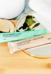 To hudplejetuber på en træoverflade: en mintgrøn 25ml "Hyaluronic Acid Power Essence" og en beige 15g "Good to Glow - Gold Mask."