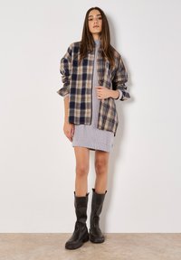 Chemise à carreaux en flanelle à boutonnage dans les tons bleu marine et beige, portée par-dessus une robe grise à côtes. Des bottes noires montantes jusqu'aux genoux complètent la tenue.