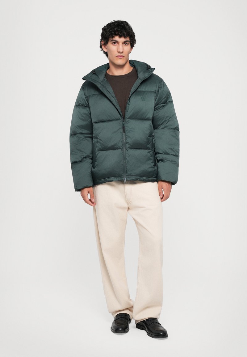 Junger Mann steht in einer dunkelgrünen Puffjacke, braunem Hemd, cremefarbenen weiten Hosen und schwarzen Slippers vor einem weißen Hintergrund.