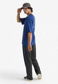 T-shirt in cotone blu con maniche corte e stampa grafica, abbinato a pantaloni grigi con strisce laterali blu. Un berretto scuro completa l'outfit.