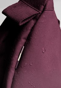 Lila wasserdichte Tasche aus strukturiertem Stoff. Verfügt über einen umklappbaren Deckel und sichtbare Wassertropfen, die ihr schützendes Design hervorheben.