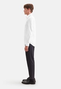 Jeune homme debout de profil portant une chemise blanche à manches longues, un pantalon noir et des chaussures habillées noires sur un fond clair uni.