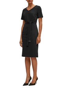 Schwarzes Kleid mit V-Ausschnitt, kurzen Ärmeln und einem Taillengürtel. Der Stoff ist glatt, und der Saum endet knielang. Kombiniert mit schwarzen spitz zulaufenden Absätzen.