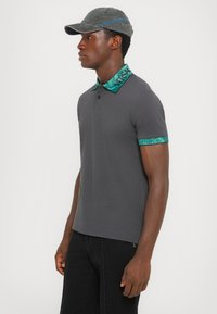 Grå polo-skjorte med en teksturert turkis krage og mansjetter. Følger med en matchende caps med blå tekst. Minimalistisk design totalt sett.