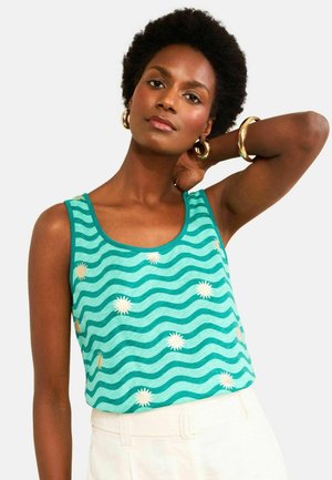 Femme aux cheveux courts et bouclés portant un débardeur bleu sarcelle à rayures ondulées et motifs de soleil blancs, un pantalon blanc, des boucles d'oreilles créoles dorées et un bracelet.