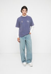 Violeta kokvilnas t-kreklu kārtota virs ilgiem piedurknēm baltā krekla, saskaņota ar gaiši ziliem platām biksēm un pelēku akcentu sporta apaviem.