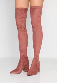 Luźne, wysokie kozaki za kolano w kolorze przykurzonego różu, wykonane z zamszu, z kwadratowym noskiem i masywnym obcasem. Widoczne szwy wzdłuż szwów.
