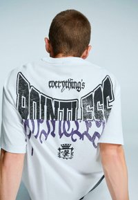 Bílý bavlněný tričko s velkým grafickým potiskem v černém a fialovém písmu na zádech. Design zahrnuje stylizované písmo a erb.