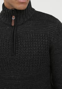 Suéter negro de punto con cuello alto, patrón texturizado y cremallera de un cuarto de longitud con un tirador de cuero marrón.