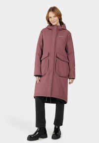 Long, pink puffer coat with a hood, featuring two large front pockets and a zip closure. Worn over black pants and ankle boots.