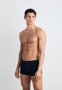 Calzoncillos boxer negros con el logo rojo "HUGO" en la cinturilla. El modelo tiene una complexión atlética, de pie con los brazos relajados y el torso visible.