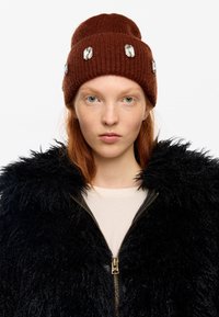 Joven con cabello rojo que lleva un gorro de punto marrón con grandes gemas y una chaqueta negra de peluche con cremallera sobre una camisa clara.