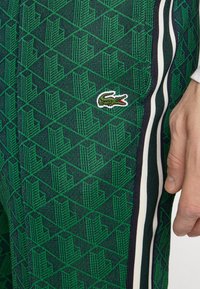 Zaļas bikses ar rakstu, kas iezīmē ģeometrisku dizainu, Lacoste logotipu un baltām sānu svītrām ar zila akcentiem, izgatavotas no teksturēta auduma.