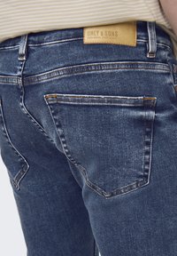Los jeans de mezclilla azul oscuro presentan un diseño ajustado con dos bolsillos traseros, costuras visibles y una etiqueta de marca de cuero en la cinturilla.