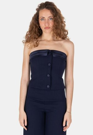Donna con capelli ricci che indossa un top blu navy senza spalline con bottoni e pantaloni a vita alta abbinati, in piedi davanti a uno sfondo bianco.