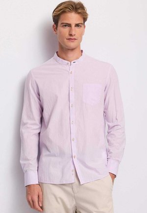 Giovane uomo in piedi con le mani in tasca, indossa una camicia viola chiaro con colletto alla coreana e pantaloni beige su uno sfondo semplice.