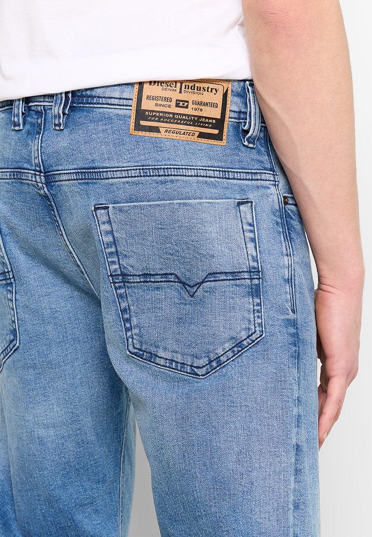 Un jean en denim bleu clair présente une coupe droite, des poches arrière avec des coutures décoratives et une patch en cuir avec logo à la taille.