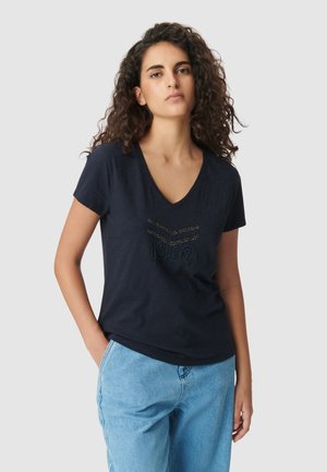 Mujer con cabello rizado vistiendo una camiseta de pico en color azul marino con el diseño "1987" y jeans de color azul claro, de pie con una mano en el bolsillo frente a un fondo gris.